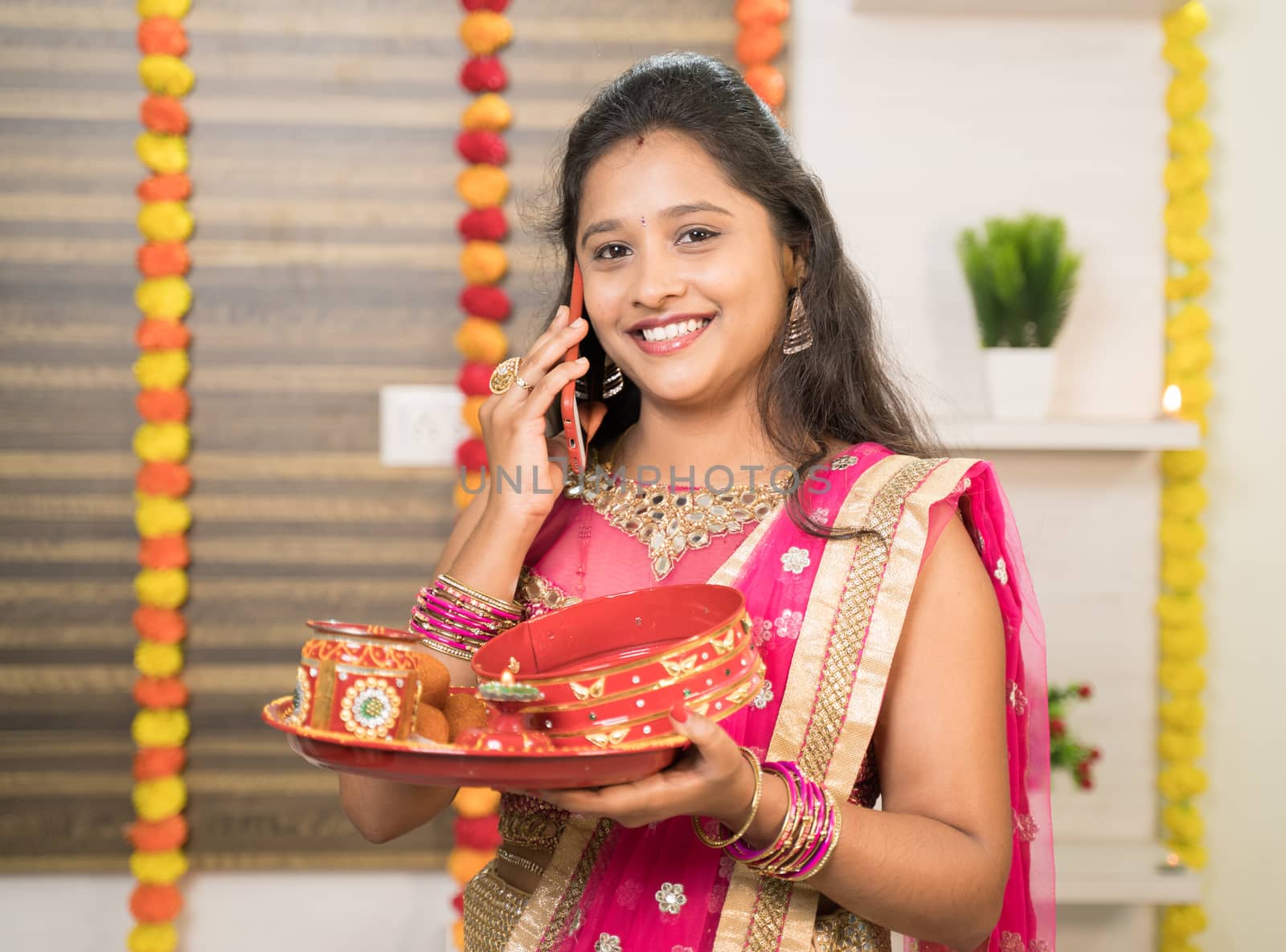 Smiling Indian Woman in traditional dress holding Karva Chauth Thali or plate while busy in talking on mobile