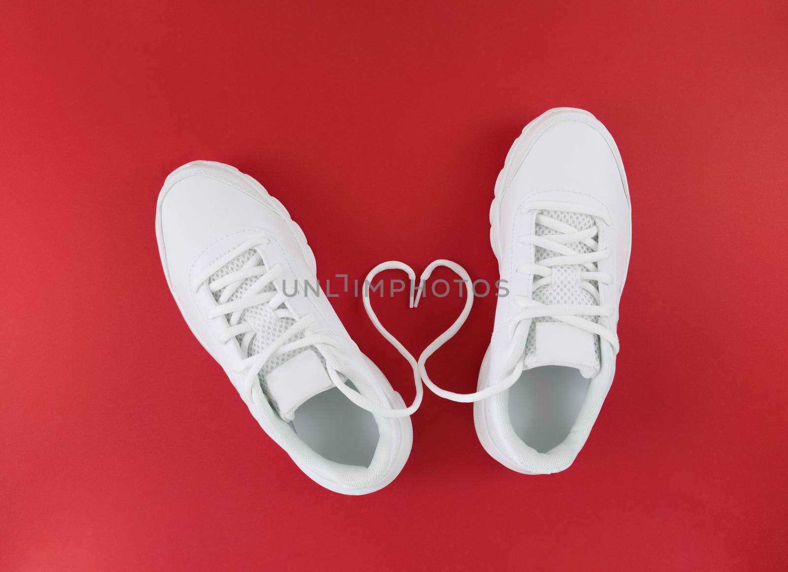 White sports shoes and heart shape from laces on red background. Simple flat lay.