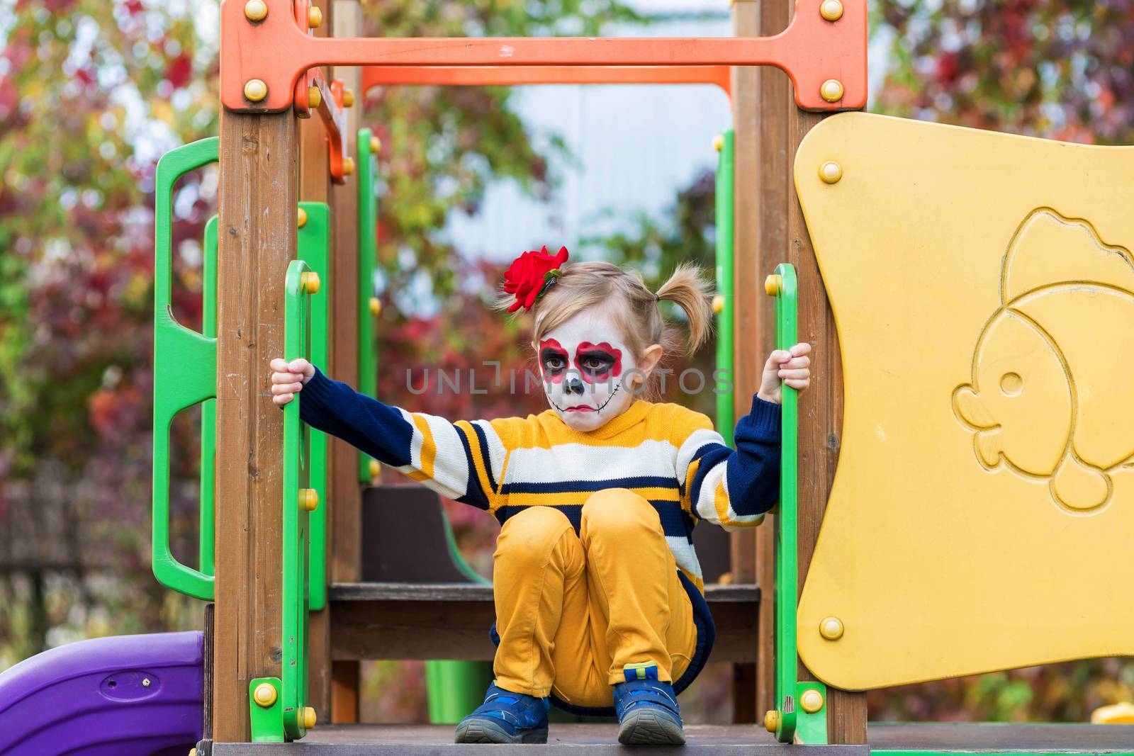 A little girl with Painted Face, rides a slide on playground on Day of the Dead. by galinasharapova