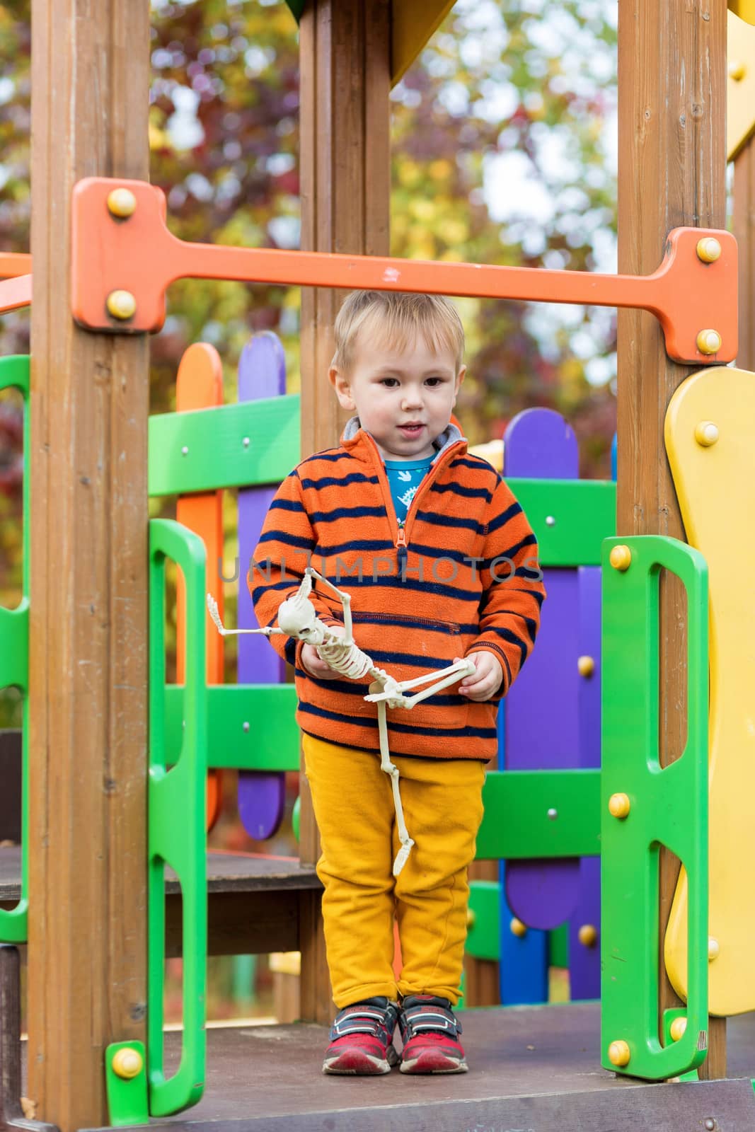 A small child in a striped sweater holds a skeleton toy in his hands and rides down a slide on the playground