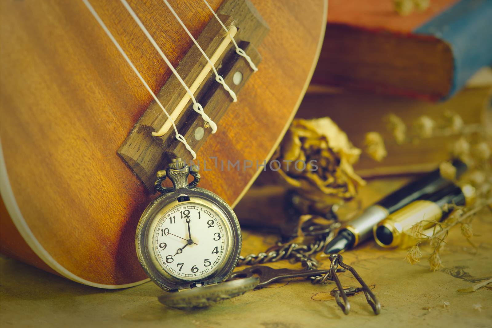 An antique pocket watch leaned against a ukulele and old book with vintage map and brass pen placed on wooden table. closeup and copy space for text. The concept of memories or things in the past.