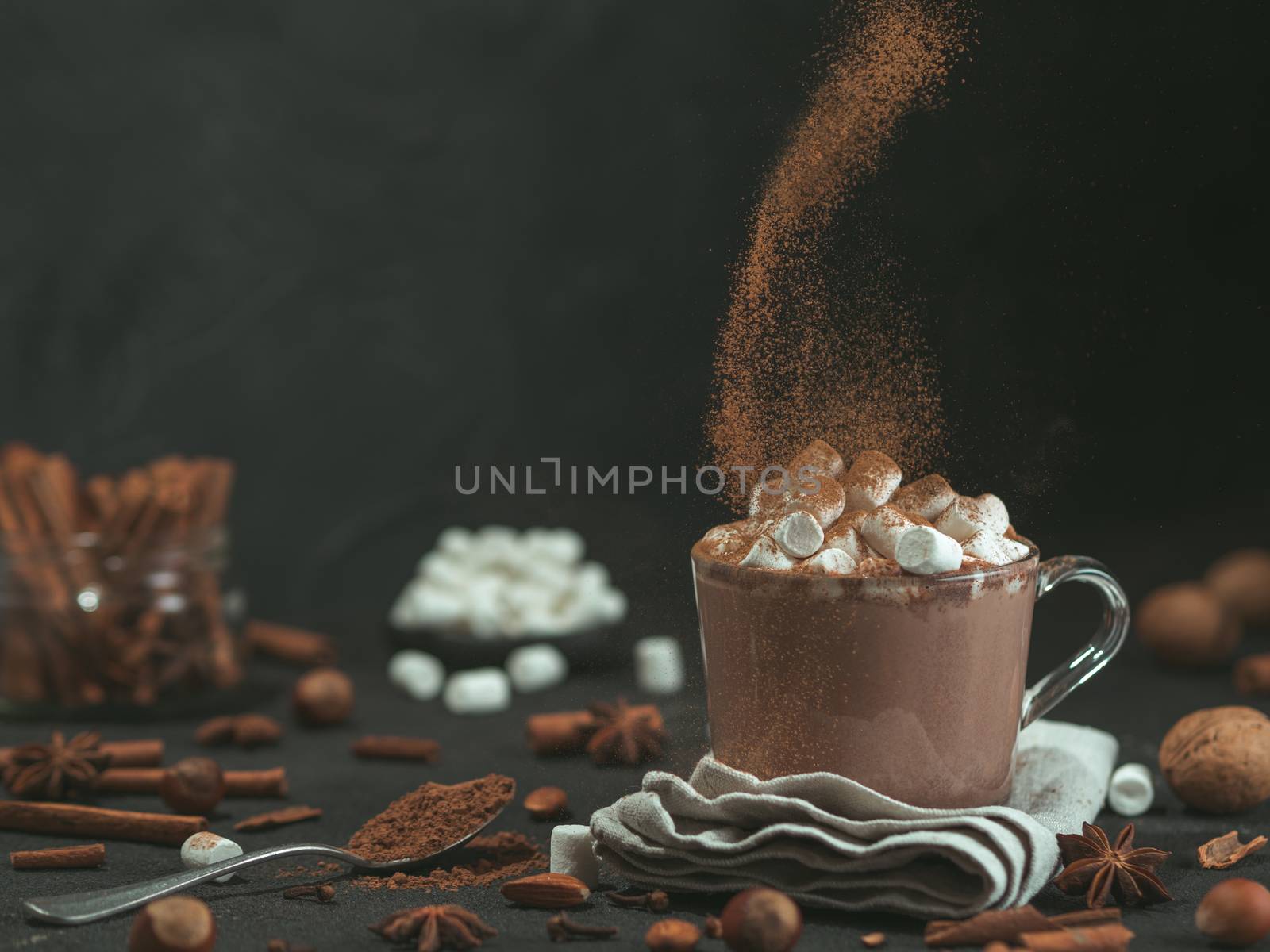 Hand sprinkled cinnamon powder on glass mug with hot chocolate cocoa drink. Copy space. Dark background. Low key. Winter food and drink concept.