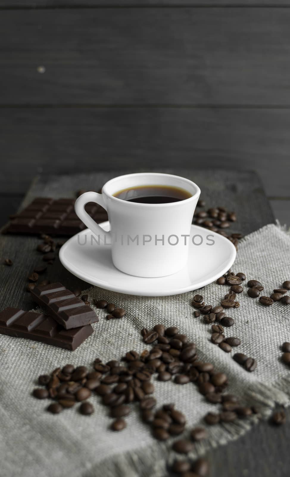 Coffee cup with cookies and chocolate with scattered coffee beans on linen and wooden table background. Mug of black coffee