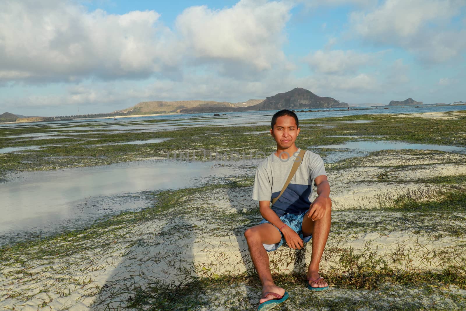 Relaxed young man meditating while sitting on the beach. Portrait of an attractive young man on a tropical beach. Teenager in a blue-gray T-shirt sitting on the beach.