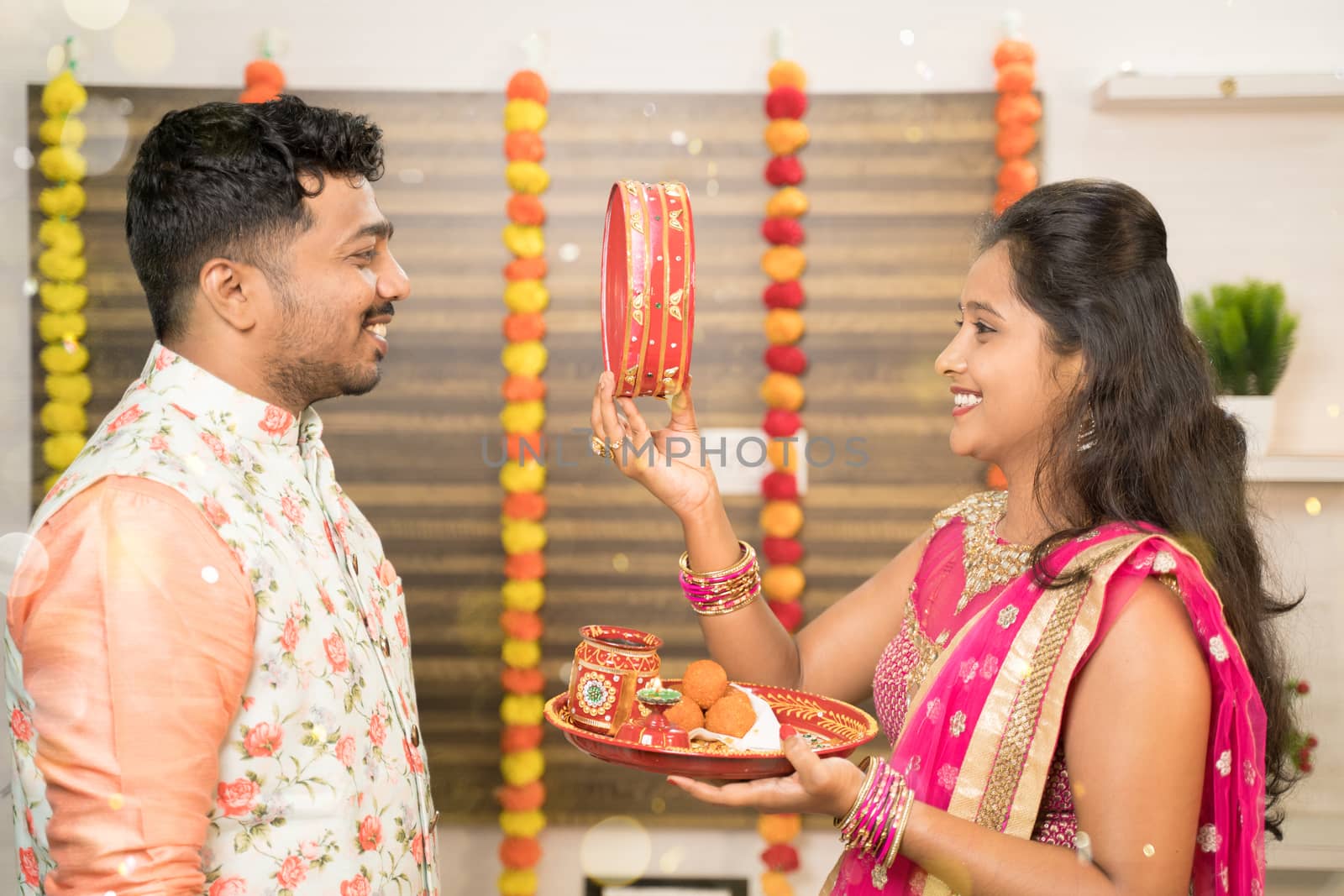 Indian woman in traditional dress seeing her husband or partner through sieve during Hindu Indian religious karwa chauth festival at home. by lakshmiprasad.maski@gmai.com