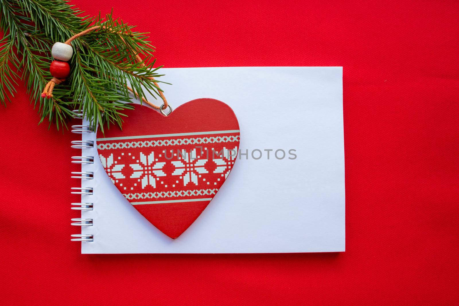 Festive composition Notepad, red wooden heart with white ornament and green fir branches on a red background.Christmas and Valentine's day concept.
