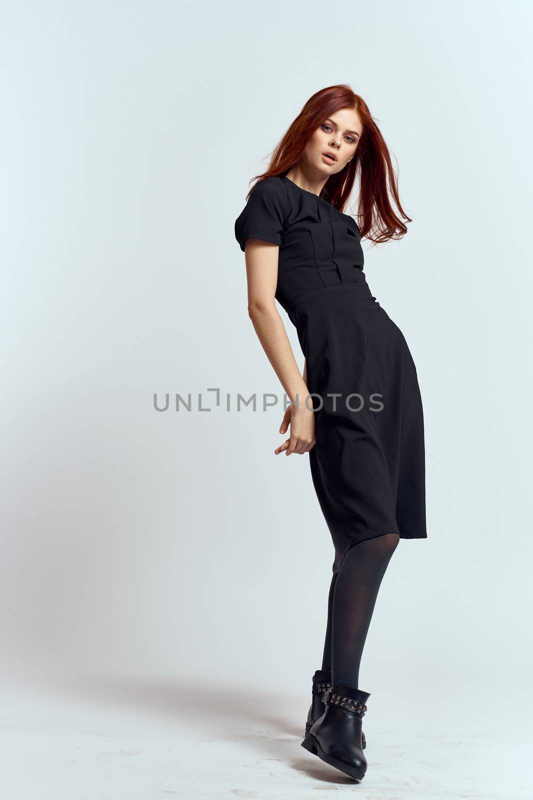 A woman in a black dress on a light background and pantyhose shoes red hair and pose in full growth by SHOTPRIME