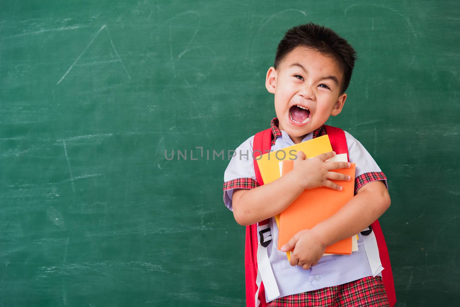 Back to School. Happy Asian funny cute little child boy from kindergarten in student uniform with school bag hold or hug books smile on green school blackboard, First time to school education concept