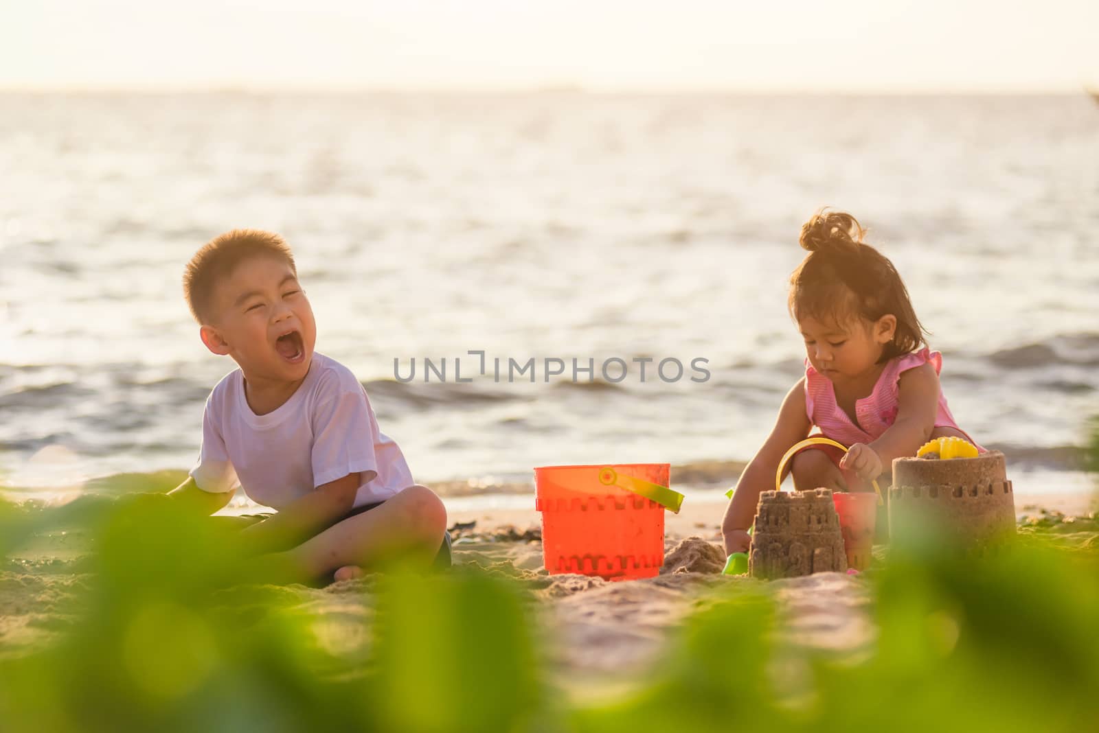 Happy fun two Asian children cute little boy and girl brother and sister family playing sand with toy sand tools at a tropical sea beach in holiday summer on sunset time, tourist trip concept