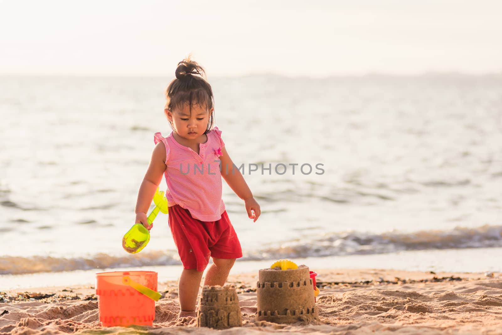 Happy fun Asian child cute little girl playing sand with toy sand tools at a tropical sea beach in holiday summer on sunset time, tourist trip concept