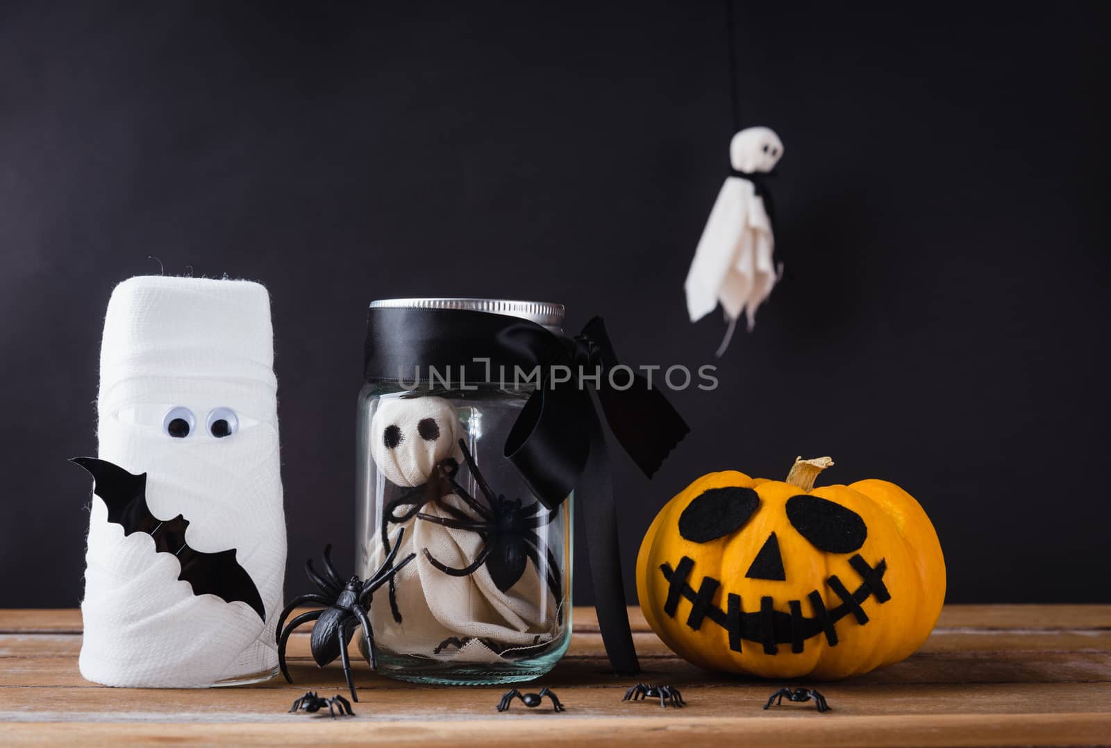 Funny Halloween day decoration party, White ghost scary face and spider in jar glass, glass of water wrapped with bandage and pumpkin on wood table, studio shot isolated on black background