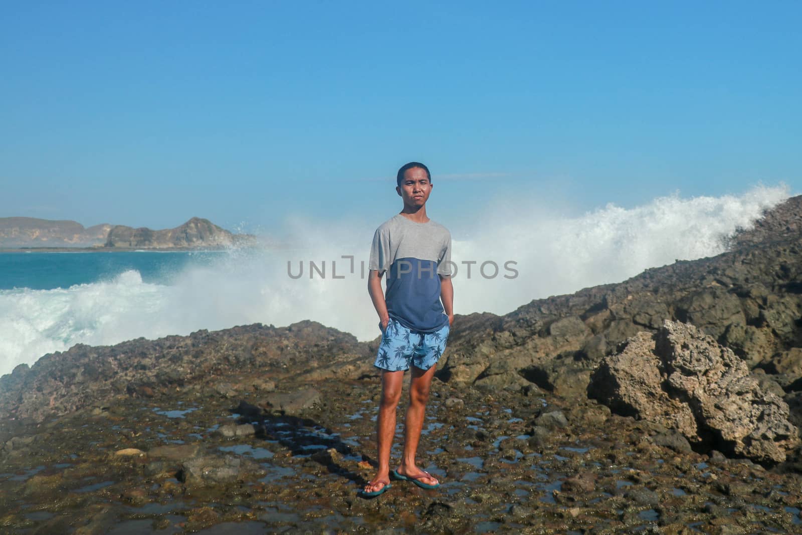 Waves hitting round rocks and splashing. A young man stands on a rocky shore and the waves crash against a cliff.