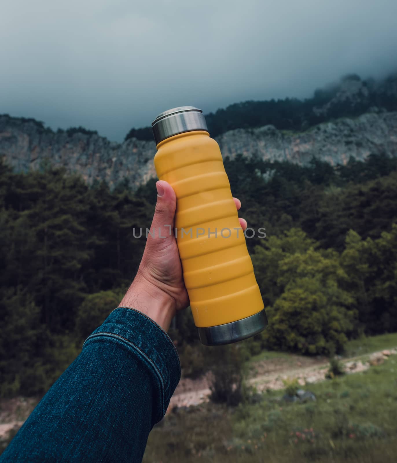 Male hand holding a steel yellow thermo bottle on nature background. by Emurado