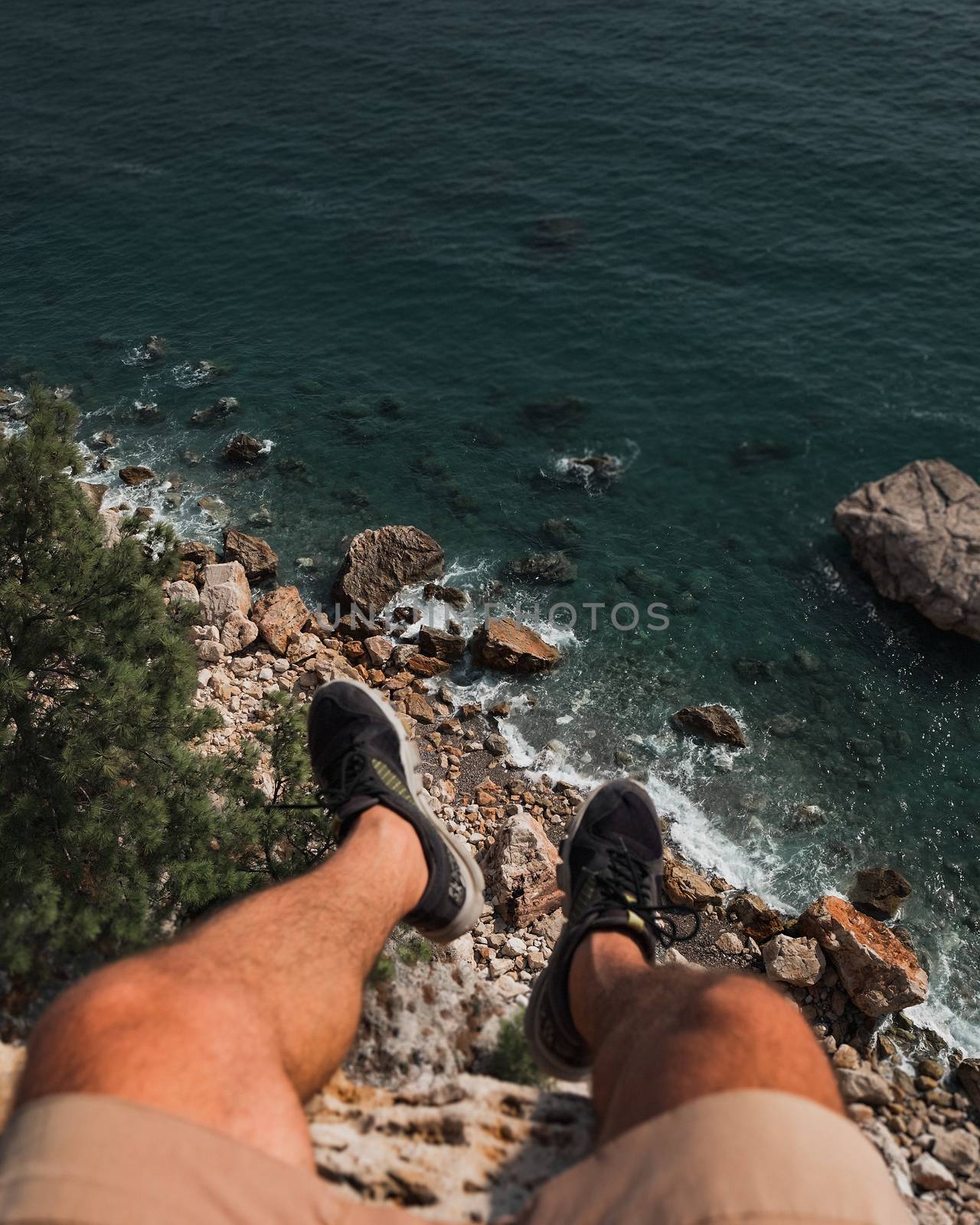 Legs of man are hanging from edge of cliff over the sea and rocks.