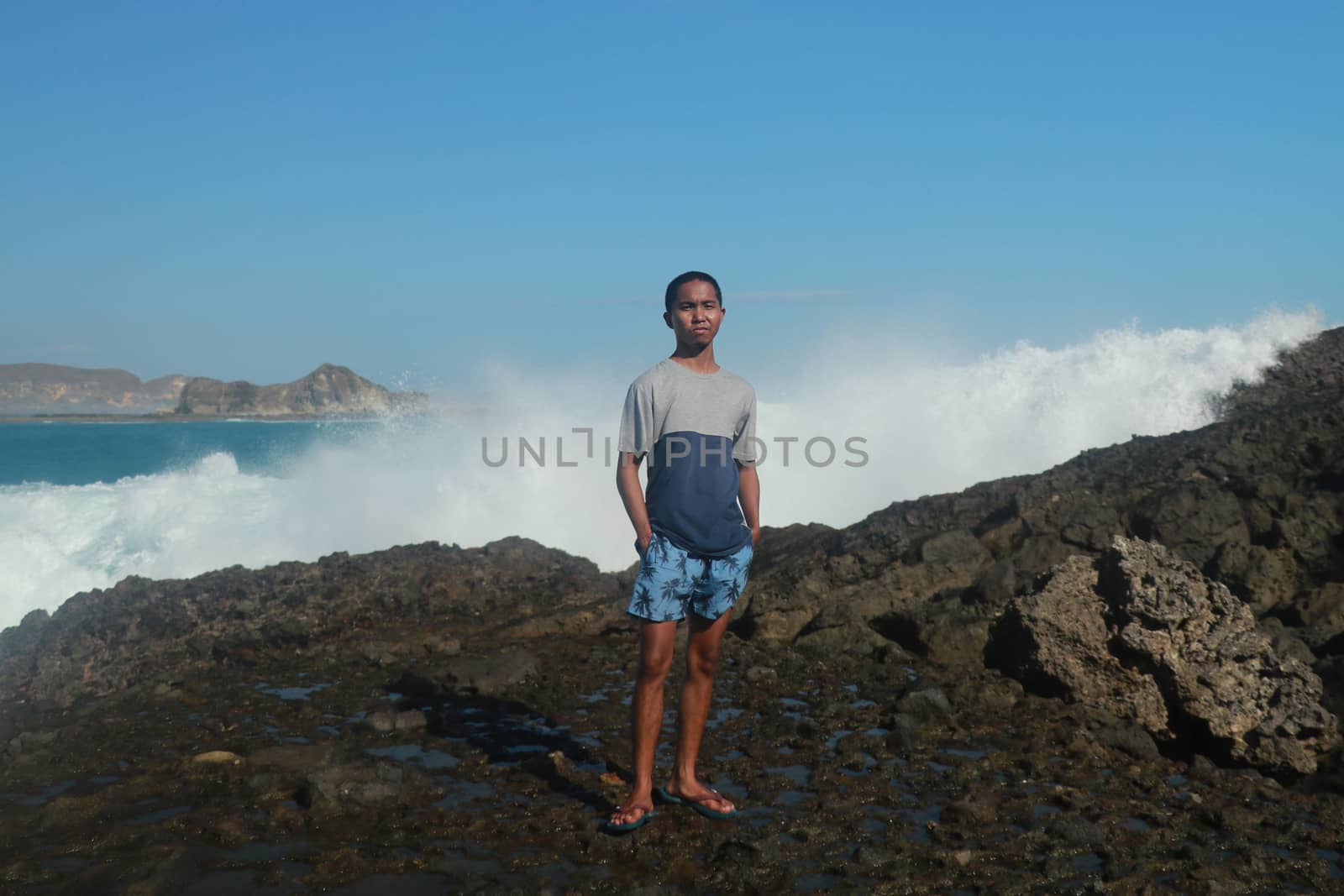 Waves hitting round rocks and splashing. A young man stands on a rocky shore and the waves crash against a cliff by Sanatana2008