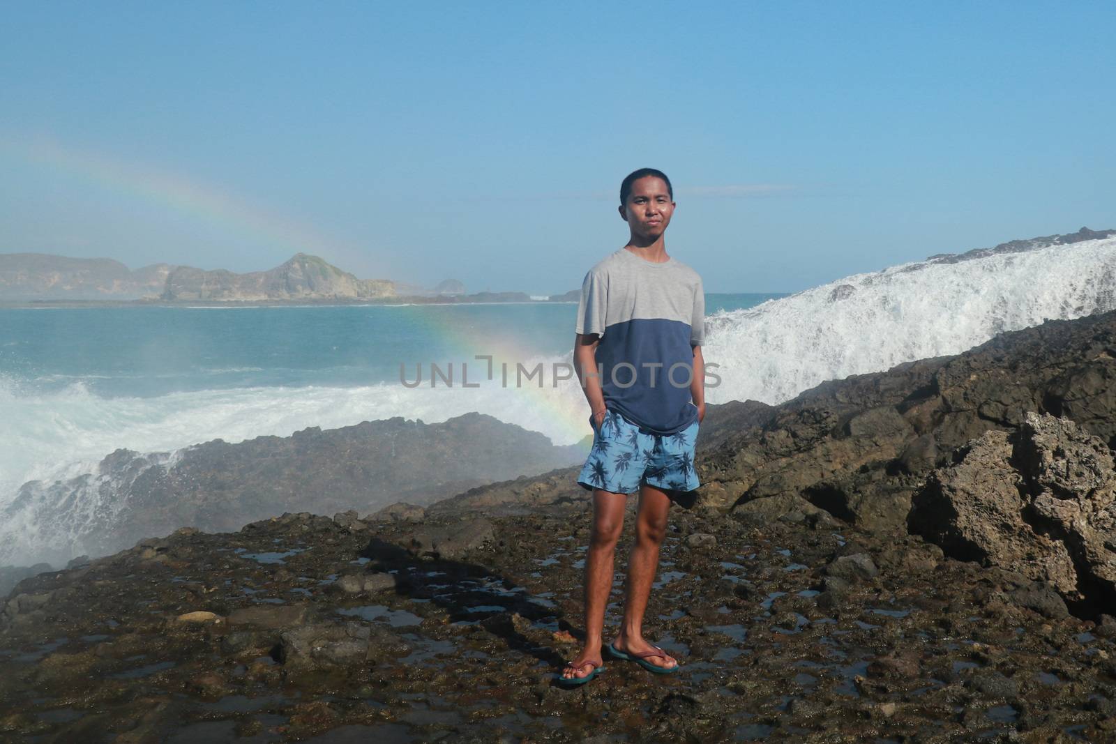 A man stands on a rocky shore and the waves crash against a cliff. Rainbow phenomenon in water fog. Waves hitting round rocks and splashing.