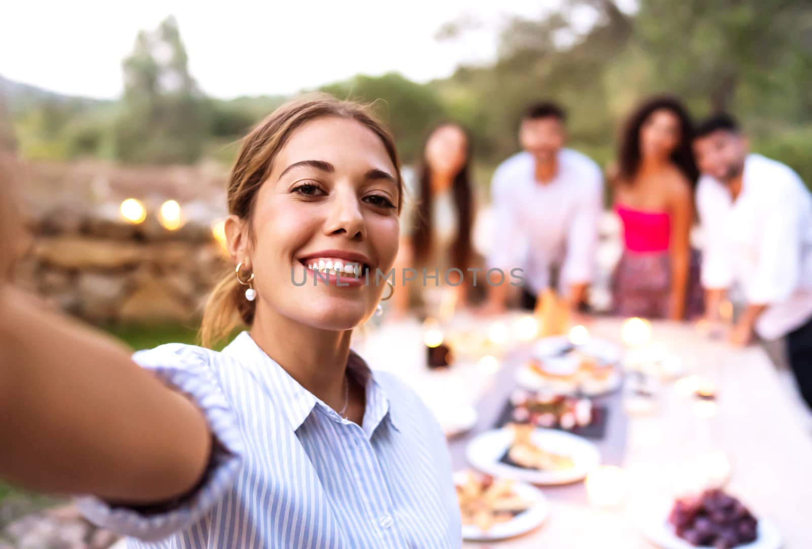 Cute smiling young woman make a self-portrait of herself and friends in the background celebrating in the garden at sunset - Happy friends moment of a home outdoor party - Female face-selective focus