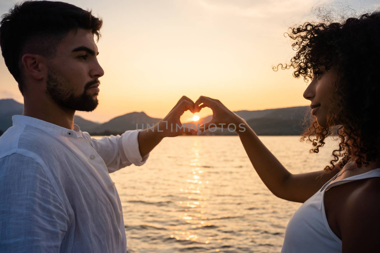 Mixed race couple, Caucasian man with her Hispanic girlfriend, forming a heart with their hands framing the setting sun on a lake water - Multiethnic lovers at sunset - Selective focus on curly woman by robbyfontanesi