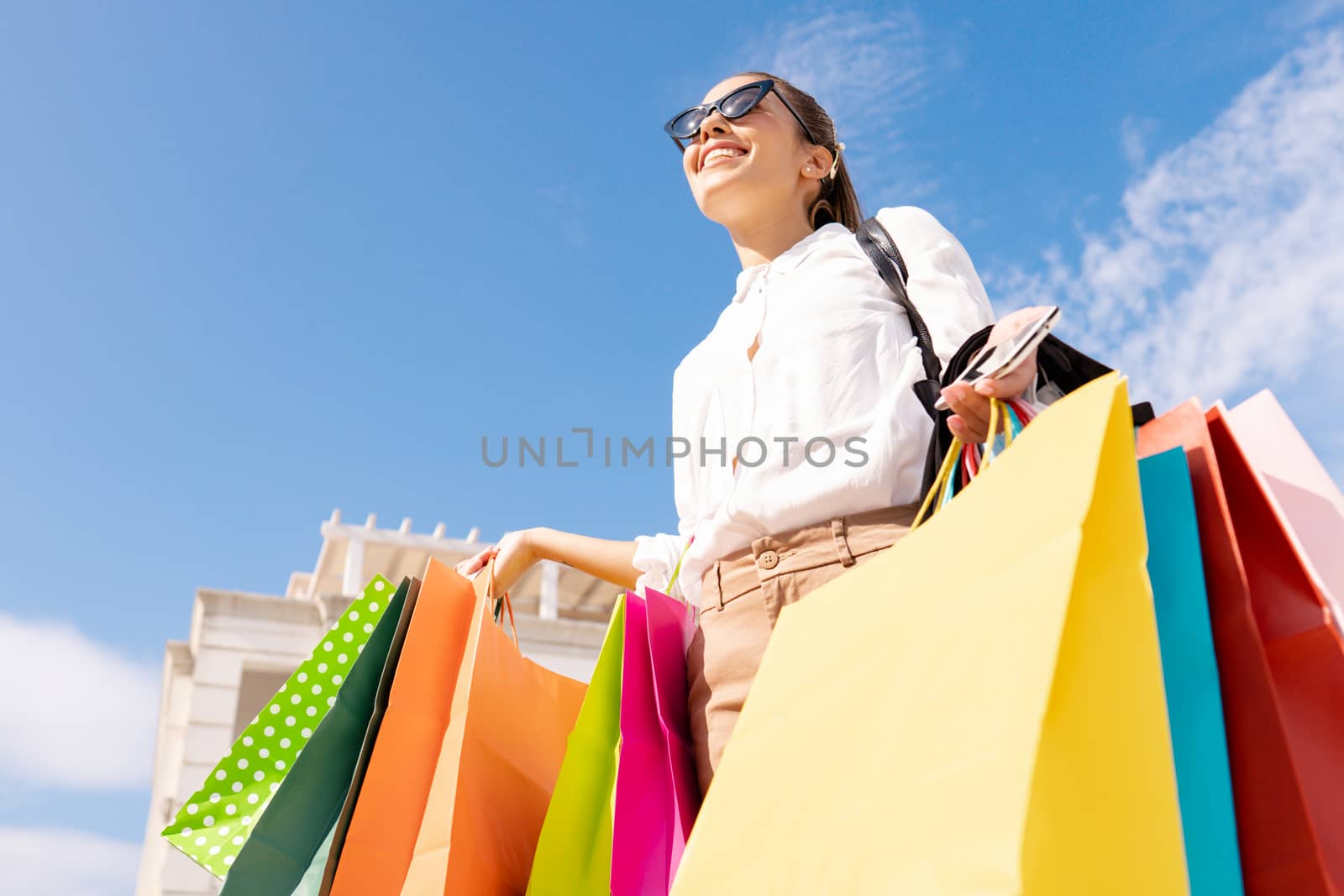 Bottom view of young beautiful stylish caucasian woman smiling holding many colorful shopping bags with blue sky background - Happy lifestyle moments of a cute female buyer by robbyfontanesi