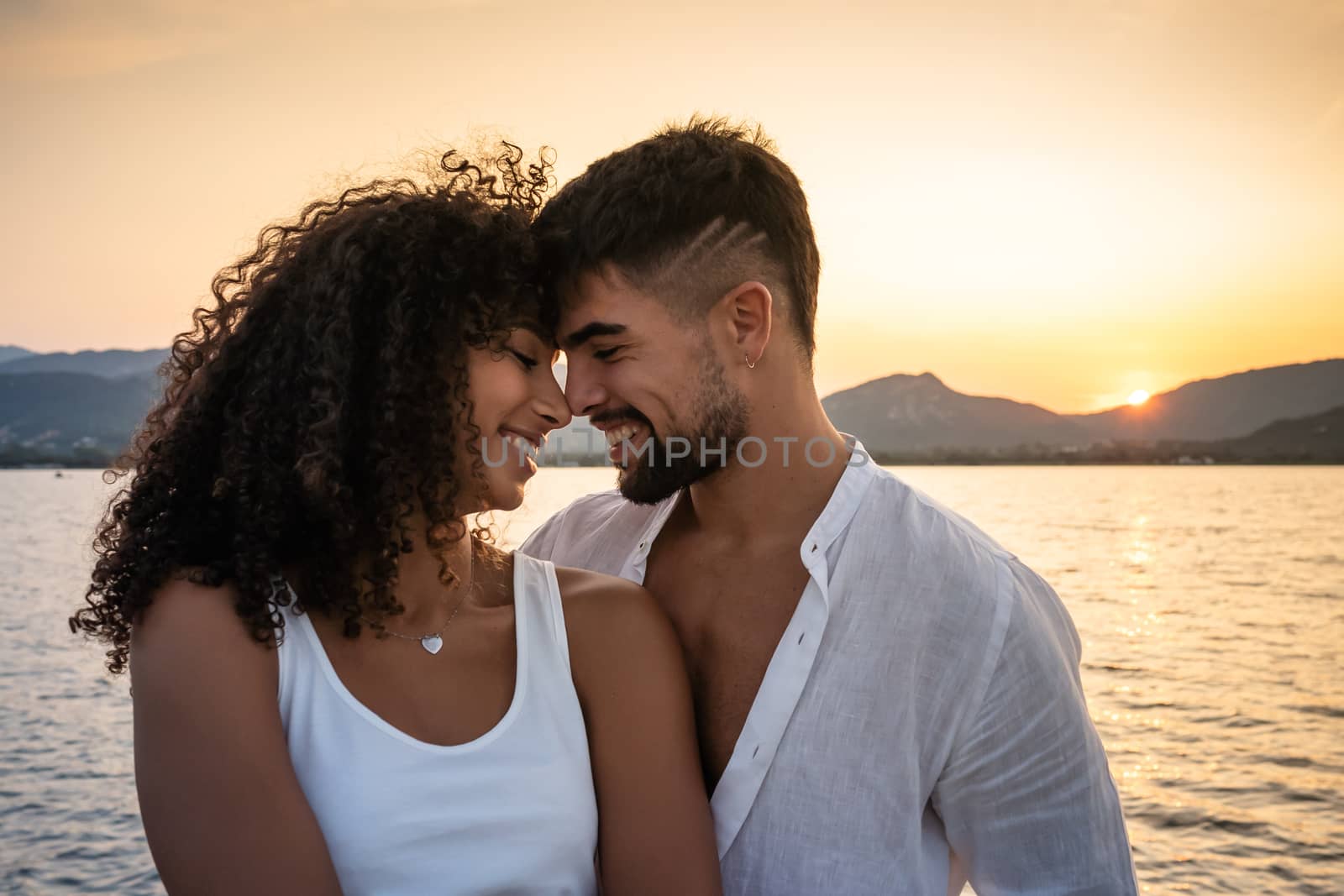 Happy mixed race couple, Caucasian attractive bearded man with her cute Hispanic girlfriend, smiling face to face looking each other with setting sun on a lake water - Multiethnic lovers at sunset
