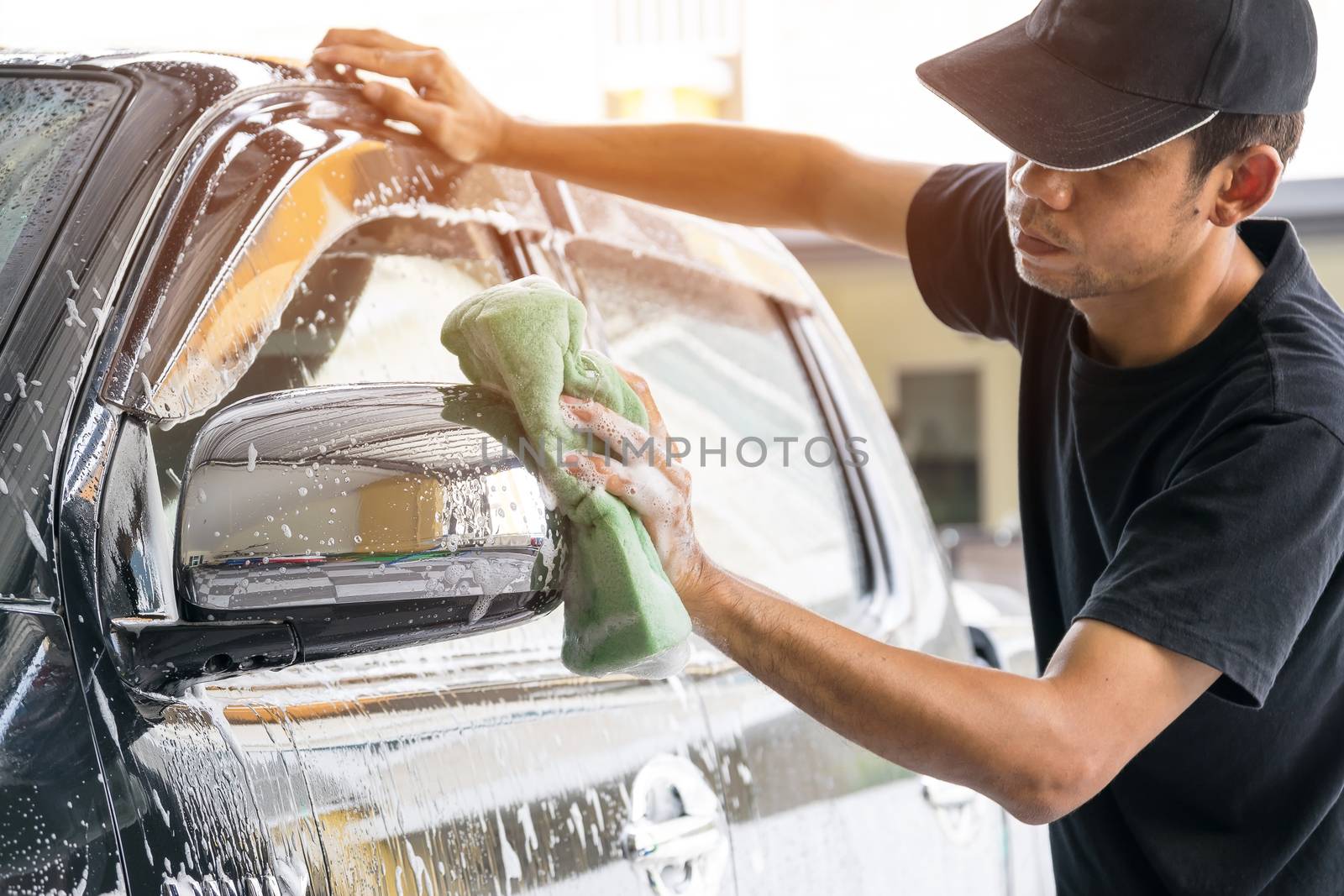 Car wash worker wearing a T-shirt and a black cap is using a sponge to clean the car in the car wash center, concept for car care industry.