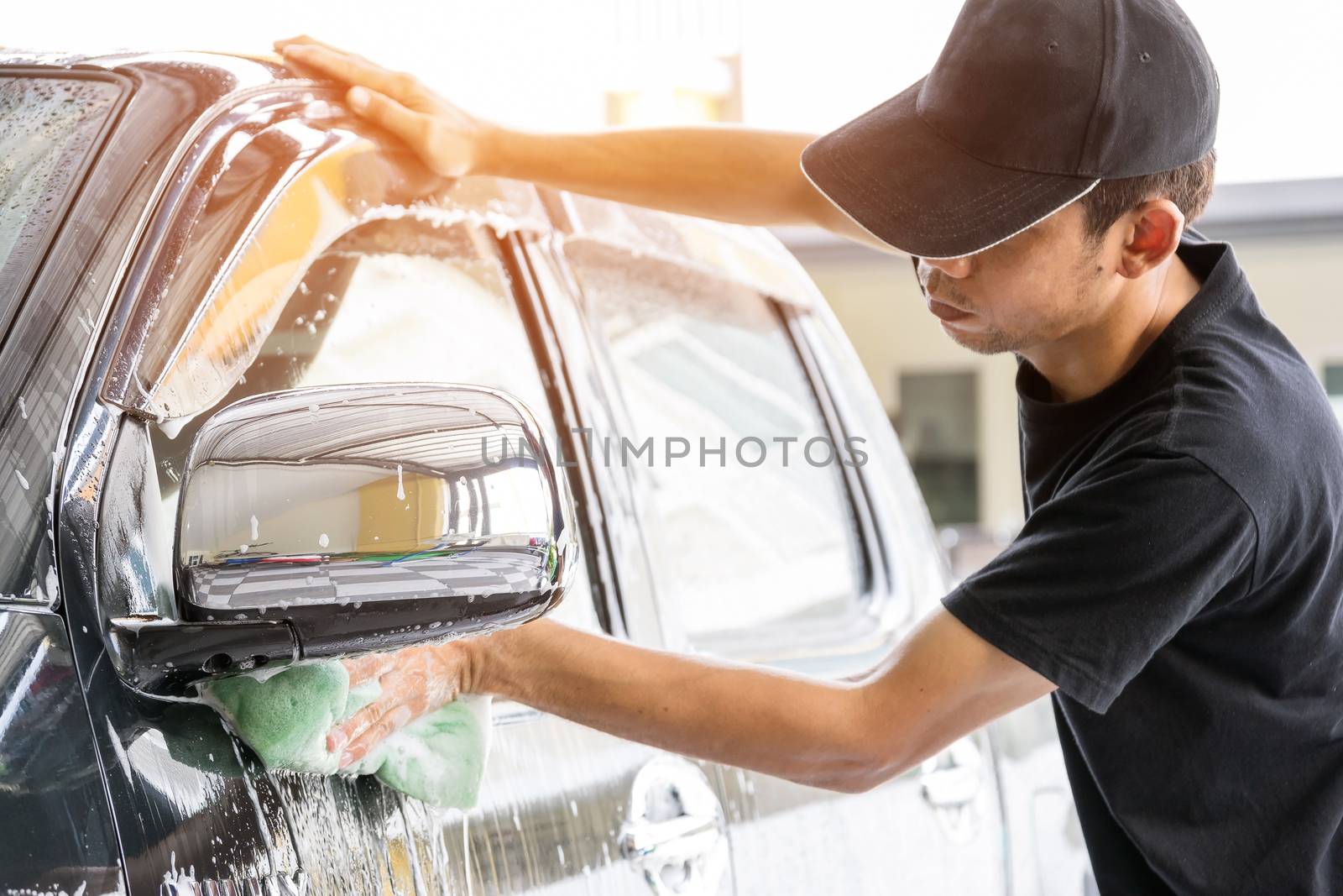 Car wash worker wearing a T-shirt and a black cap is using a sponge to clean the car in the car wash center, concept for car care industry.