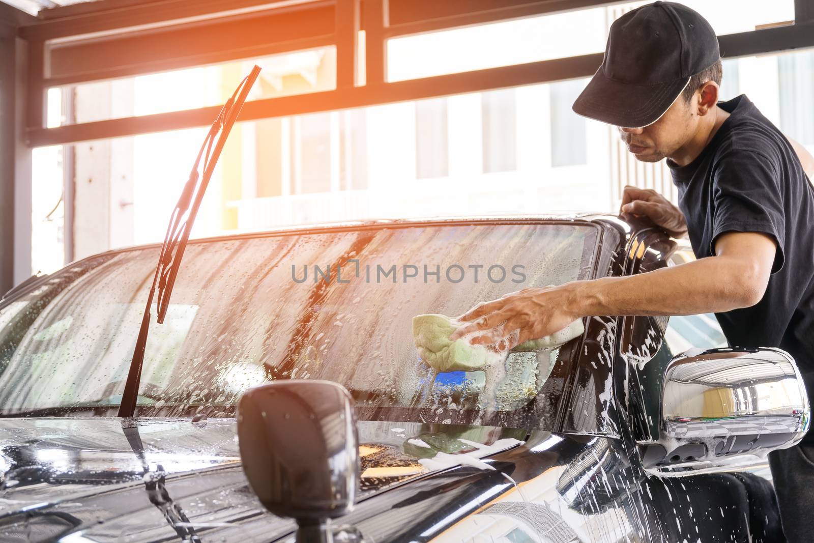 Car wash worker wearing a T-shirt and a black cap is using a sponge to clean the car in the car wash center, concept for car care industry.
