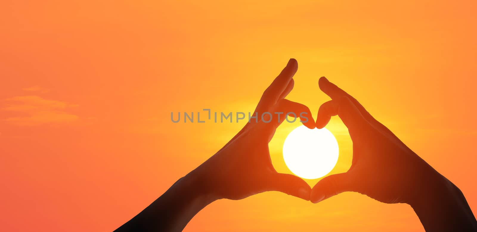 Panorama silhouette of female hands making a heart shaped symbol with beautiful sunset background.