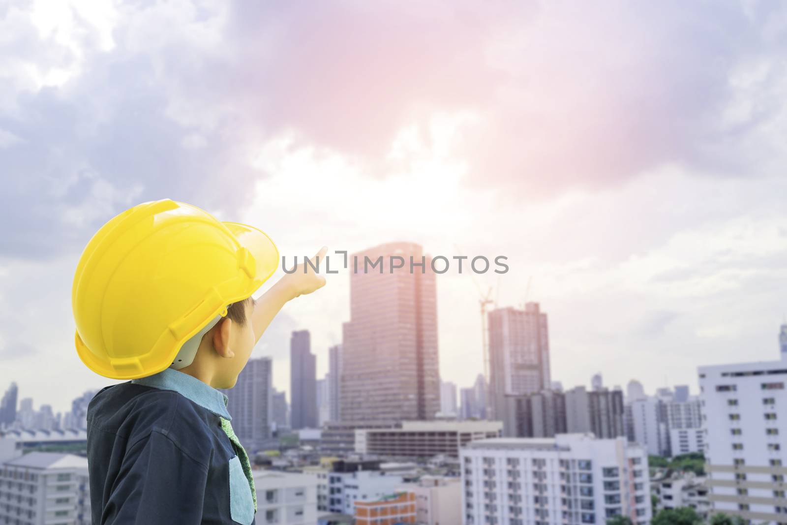 A boy dressed as an engineer wearing a safety hat pointing his hand on the cityscape with sunset background-concepts for the future is growing up to be an engineer.