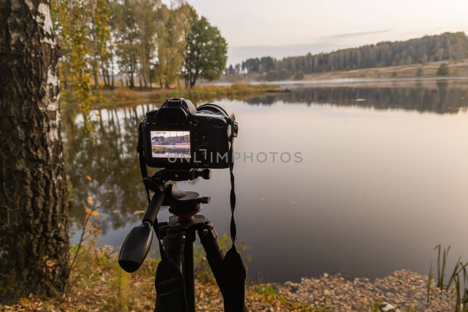 black digital camera on tripod shooting foggy early morning landscape at autumn lake with selective focus.