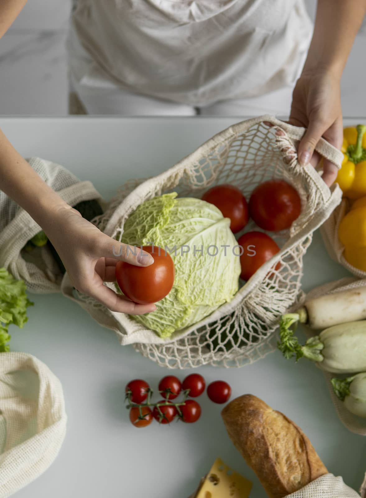 Woman came back from a market and unpacks a reusable grocery bag full of vegetables on a kitchen at home. Zero waste and plastic free concept. Girl is holding mesh cotton shopper with vegetables