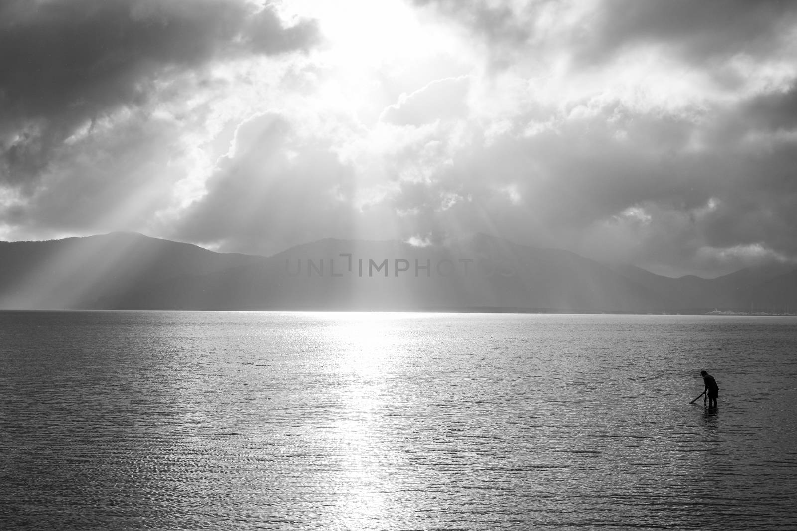 Black and white of a fisherman near the coast back-illuminated by the morning sun shining among the clouds