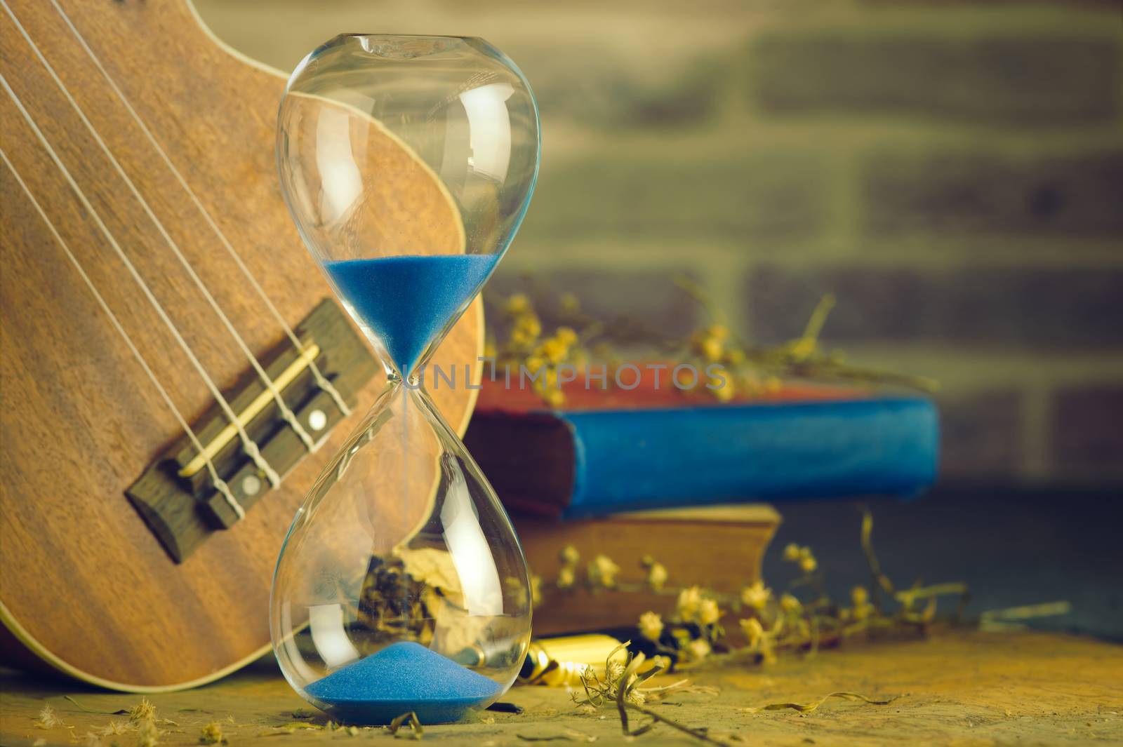 A vintage hourglass and ukulele with an old book and brass pen on a wooden table and brick background in the morning. Closeup and copy space. The concept of memories or things in the past.