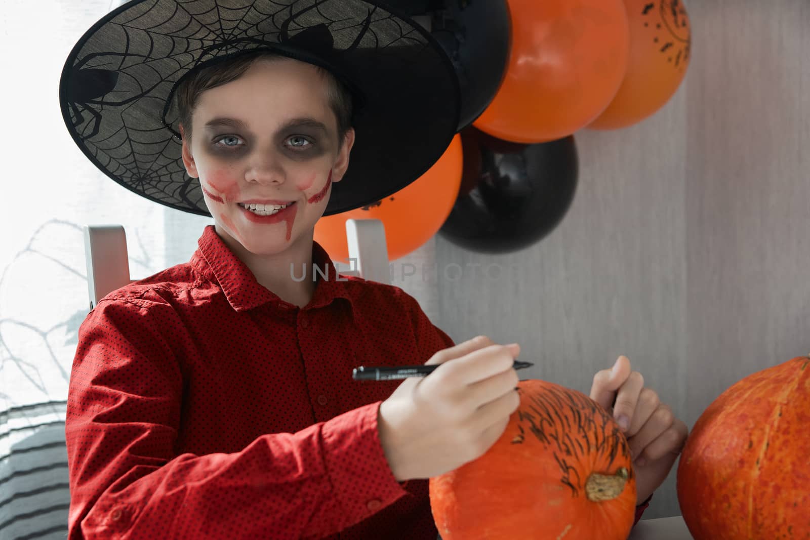 Happy teen boy in costume preparing for the Halloween celebration drawing a pumpkin. Halloween carnival or masquerade concept