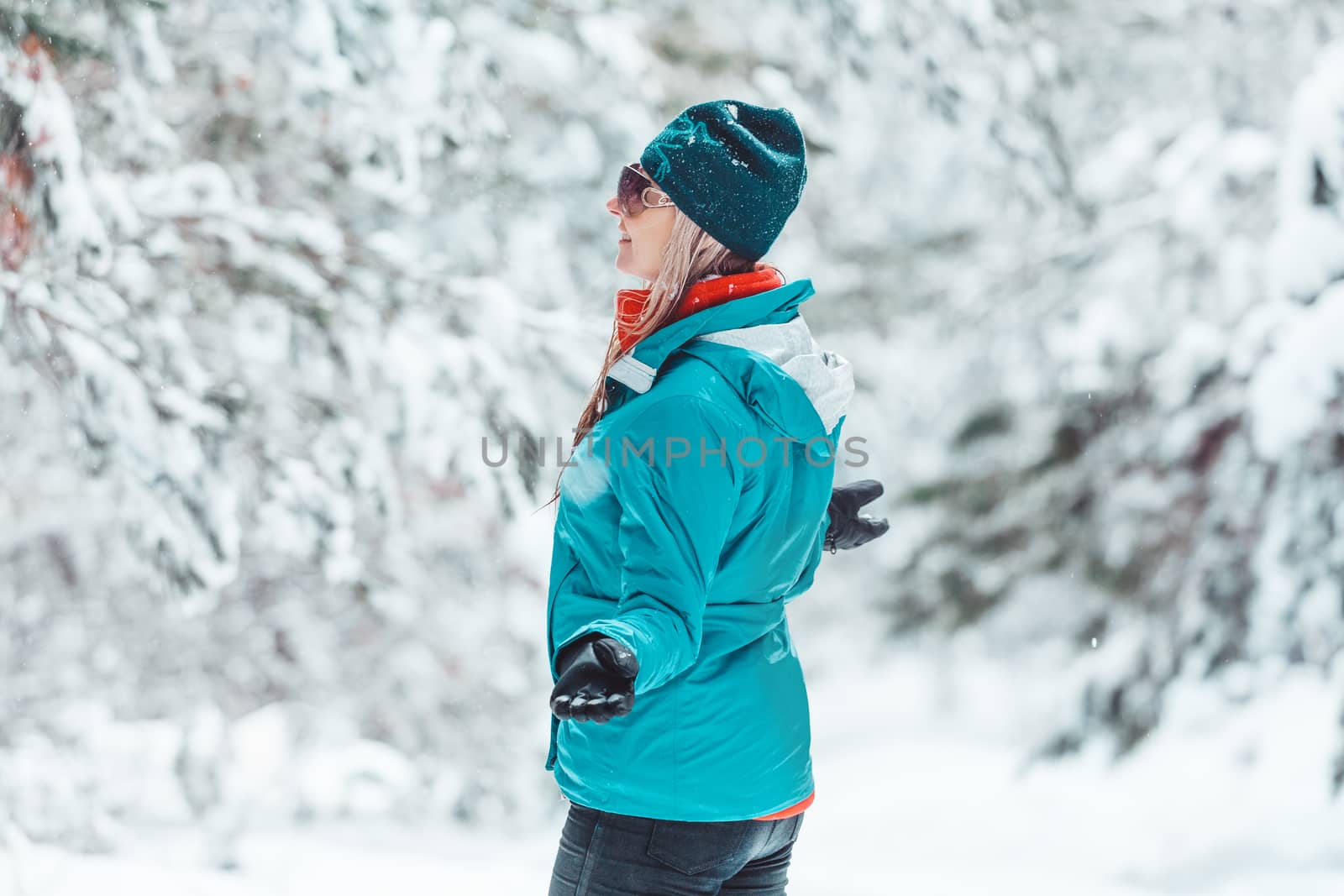 Woman wearing winter jacket and beanie standing out in falling snow  among a forest of pine trees