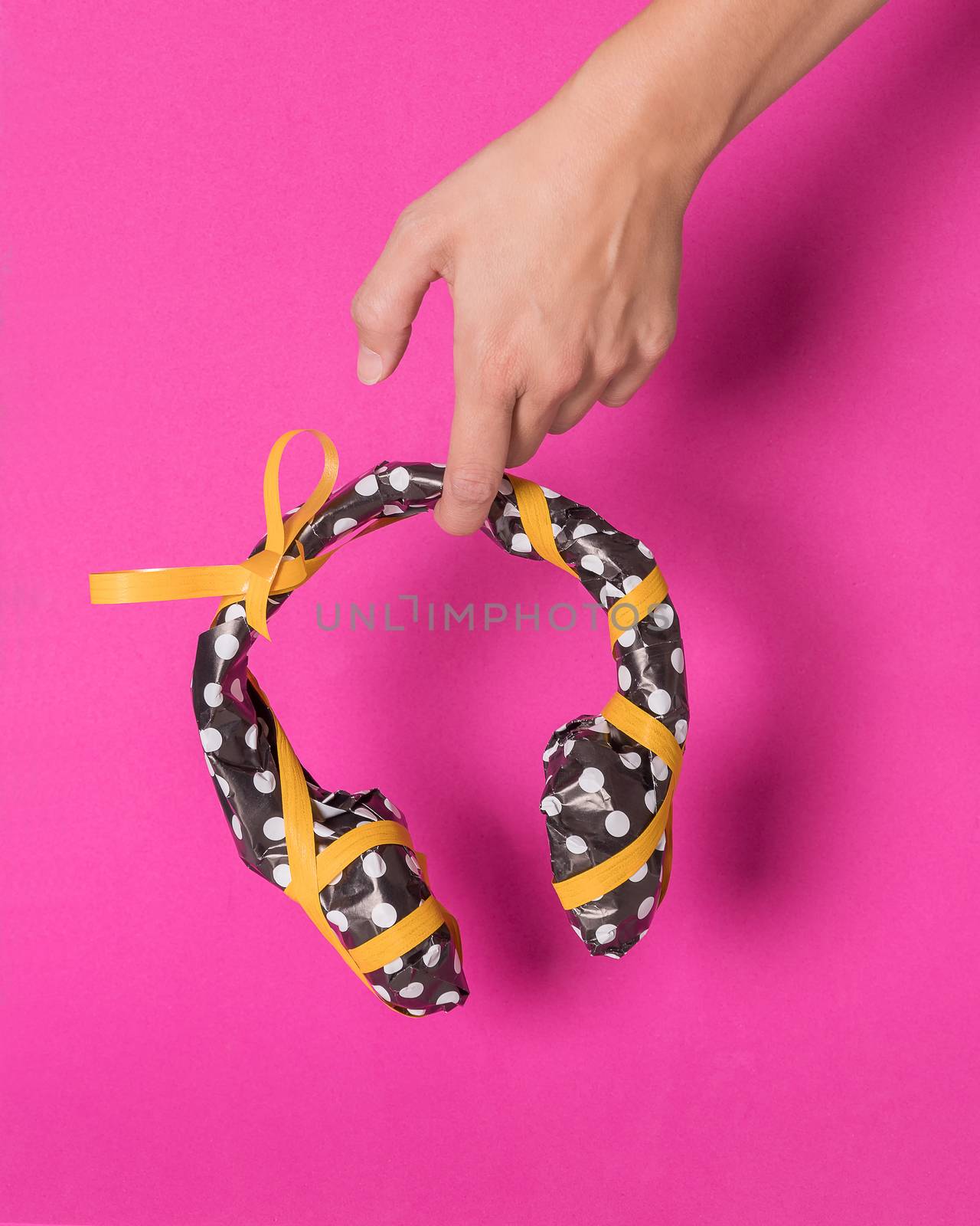 Female hand holding a headphones wrapped in black gift paper with white dots and yellow ribbon on a magenta background