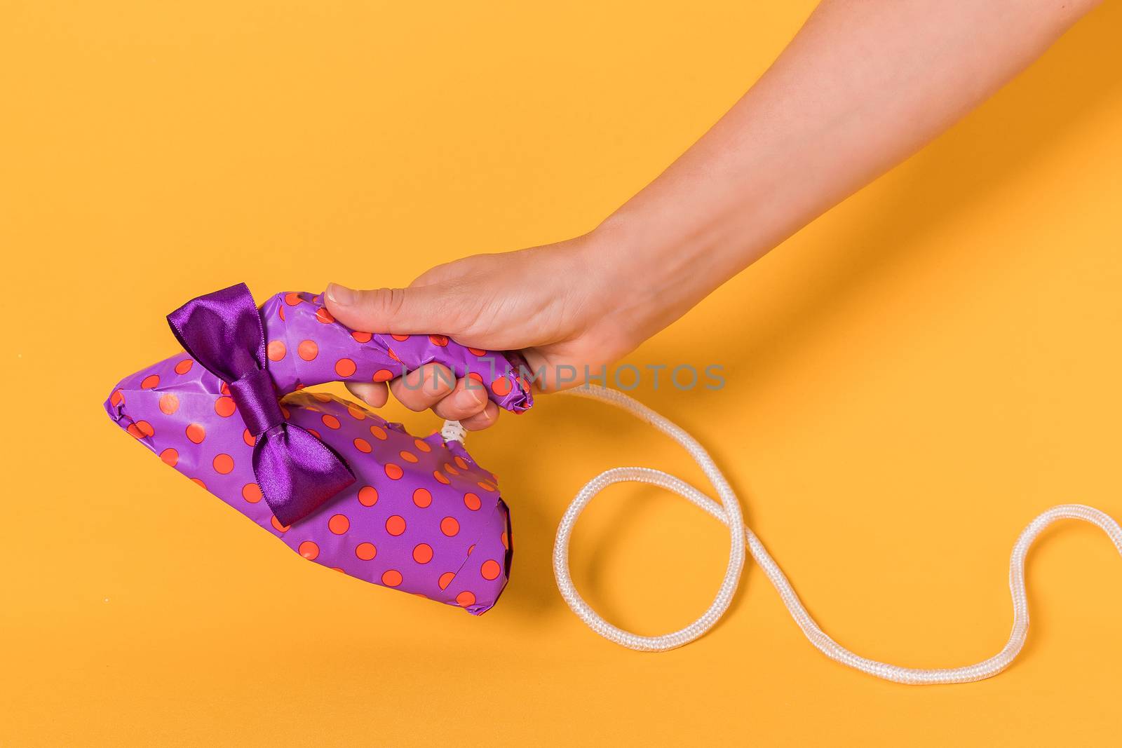 Female hand holding an iron wrapped in purple gift paper on a orange background