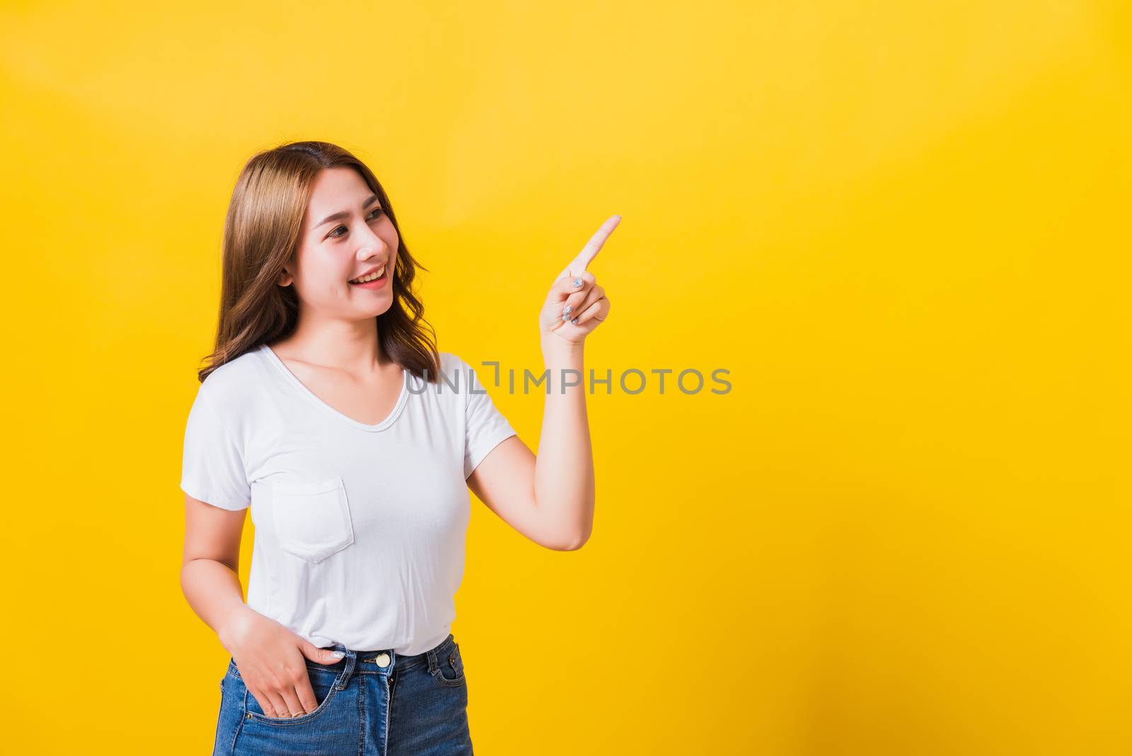 Asian Thai happy portrait beautiful cute young woman standing wear white t-shirt pointing finger away side looking to away side, studio shot isolated on yellow background with copy space