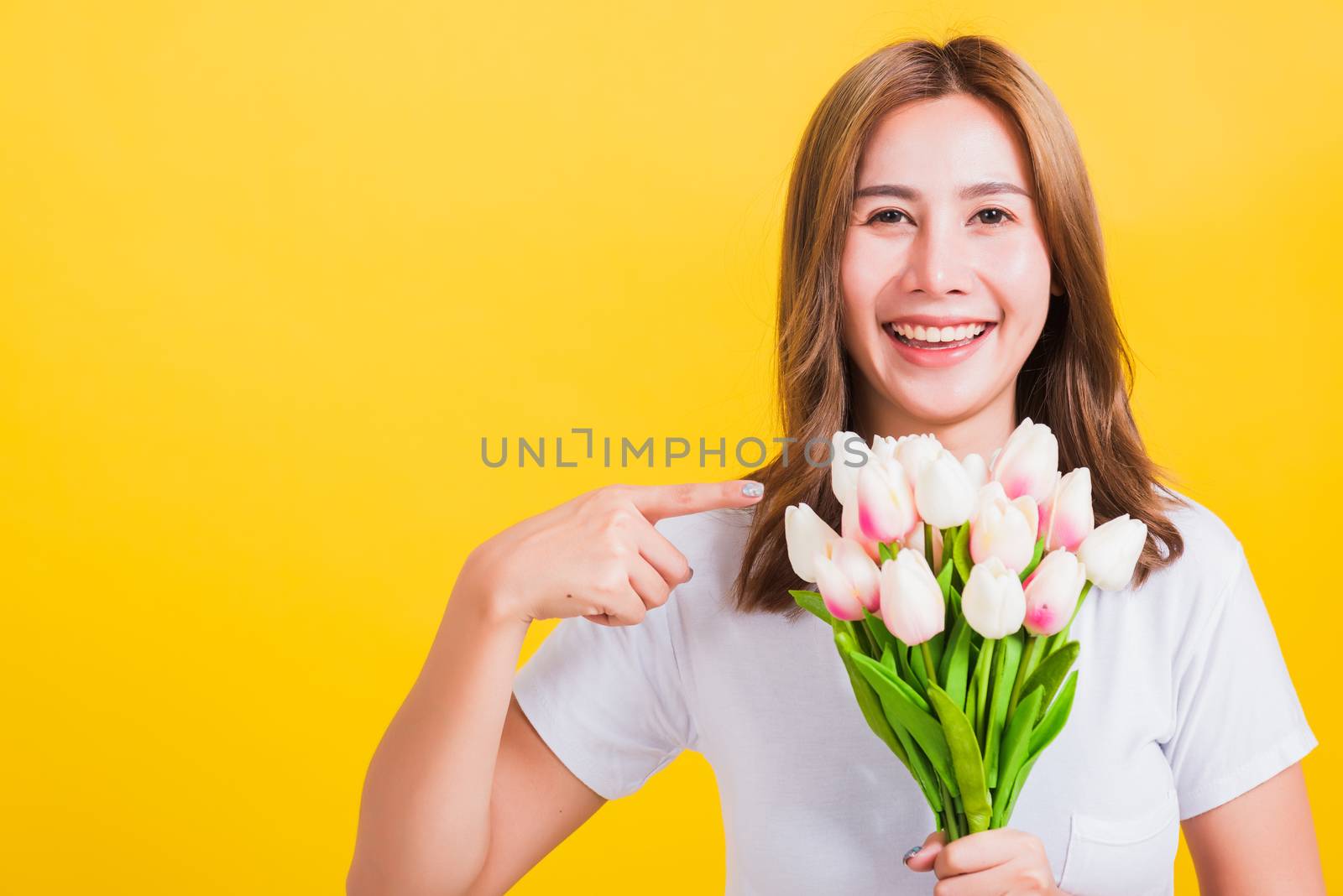 Portrait Asian Thai beautiful happy young woman smiling, screaming excited hold flowers tulips bouquet in hands and point finger to flowers, studio shot isolated on yellow background, with copy space