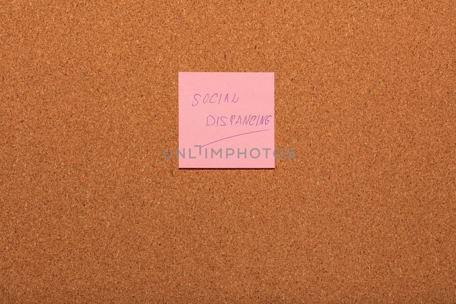 Reminder Social distancing handwritten on a pink sticker on a cork notice-board. by DamantisZ