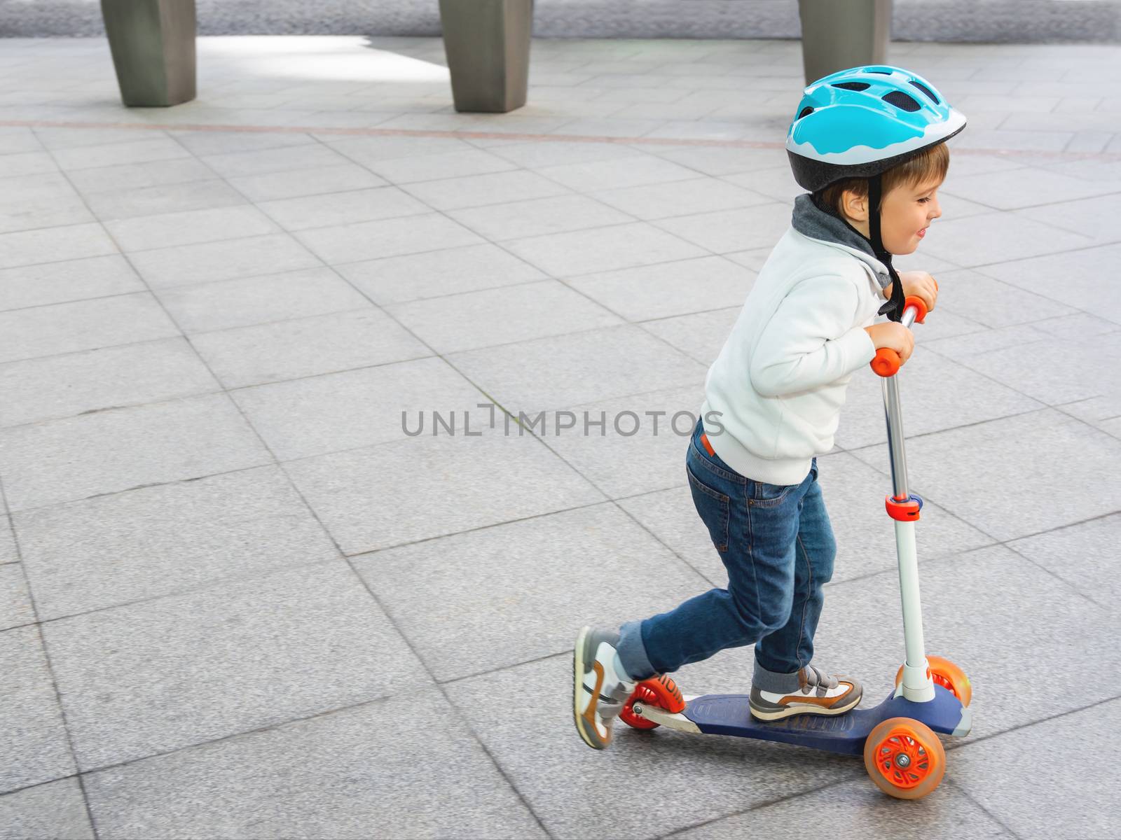 Toddler in helmet is riding scooter on parking lot with parked bicycles. Urban vehicle for active children. Leisure activity for boys and girls.
