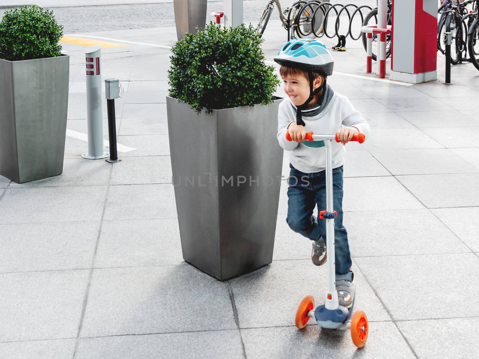 Toddler in helmet is riding scooter on parking lot with parked b by aksenovko