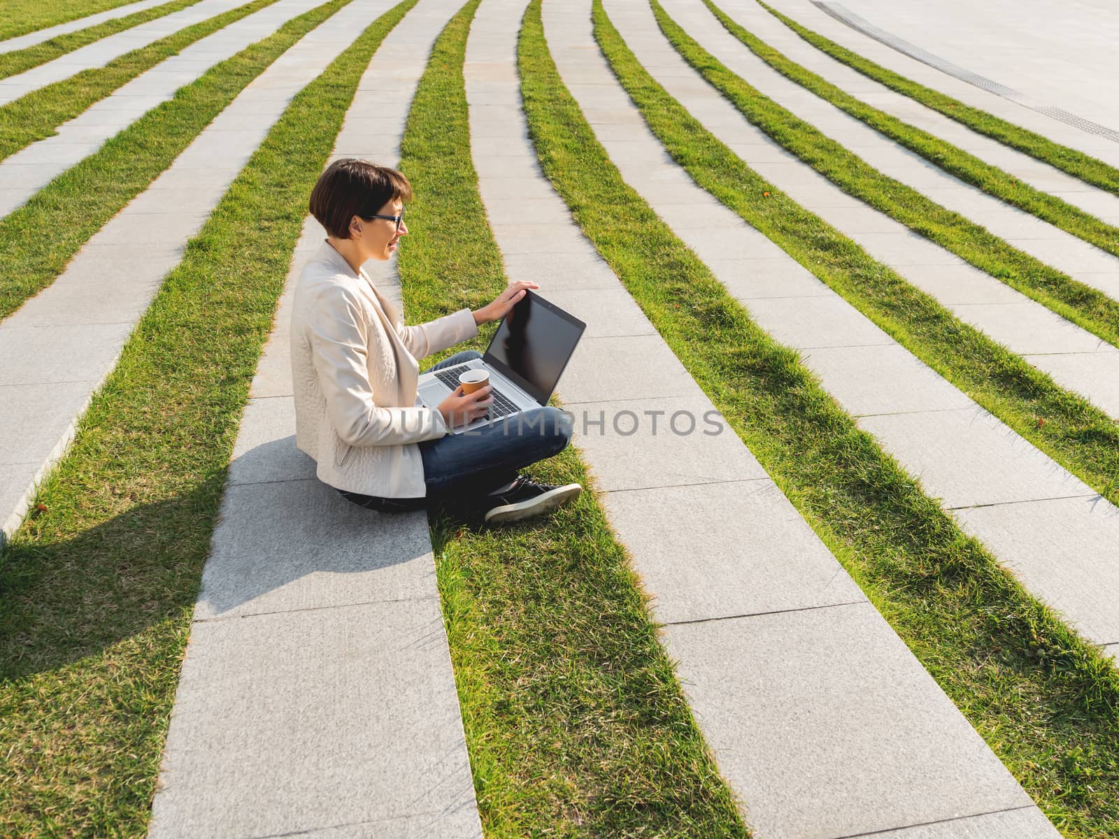 Business woman sits in park with laptop and take away cardboard by aksenovko