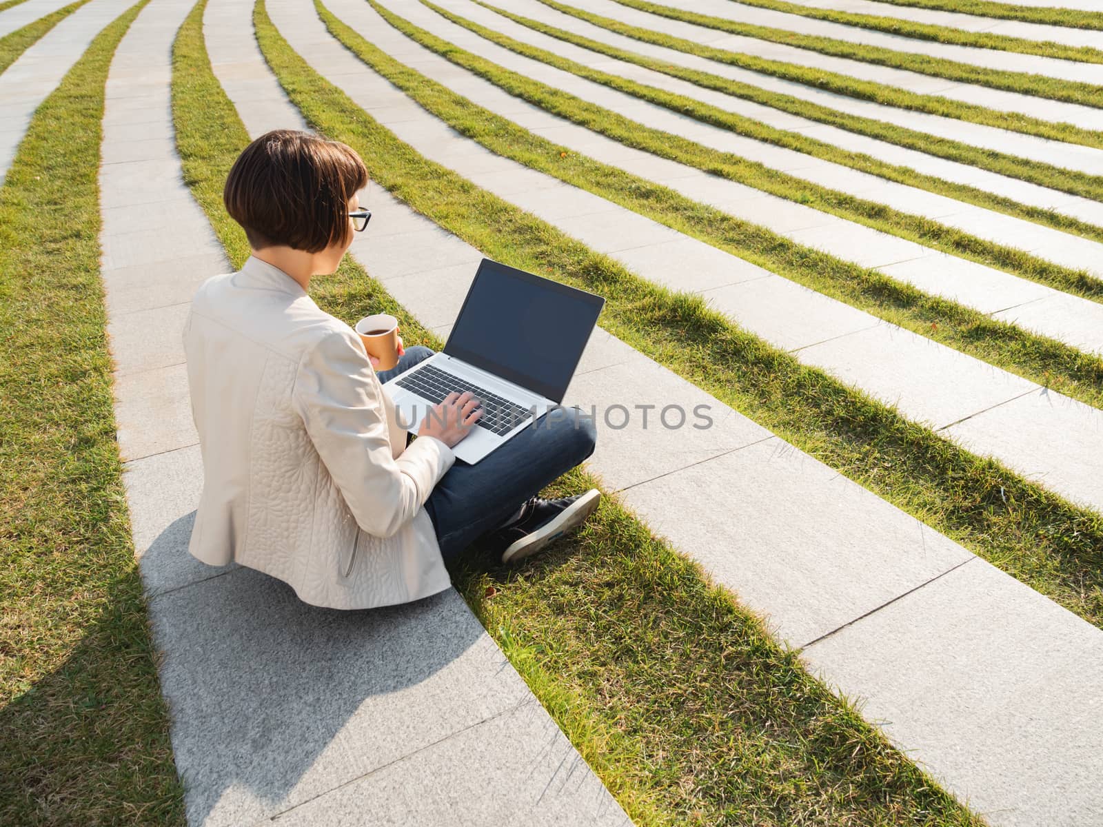 Business woman sits in park with laptop and take away cardboard cup of coffee. Casual clothes, urban lifestyle of millennials.