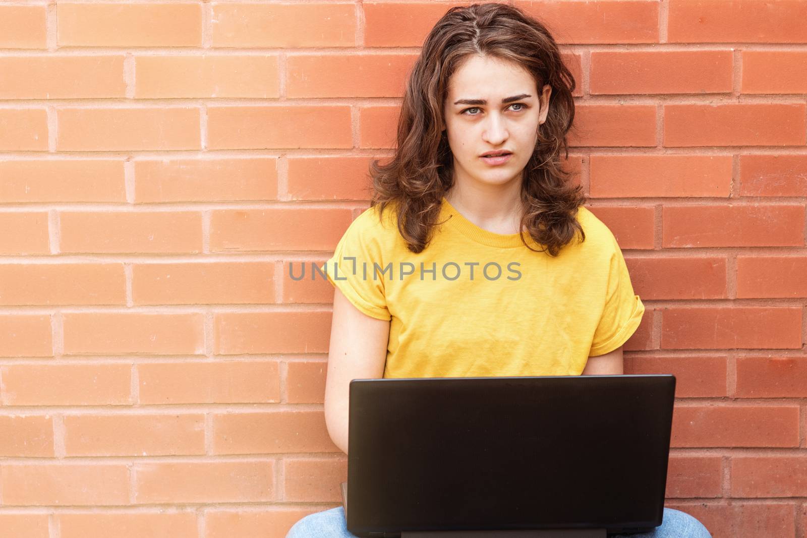 Sad young woman in yellow shirt sitting on flor, studying online and working on the laptop