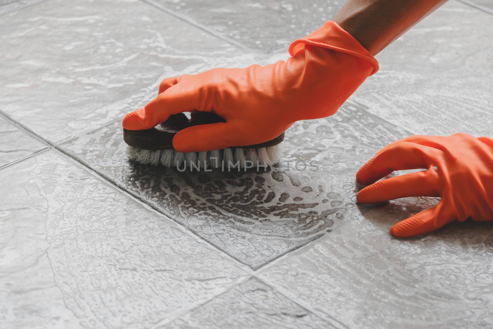 Hand of man wearing orange rubber gloves is use a hose to clean the tile floor.