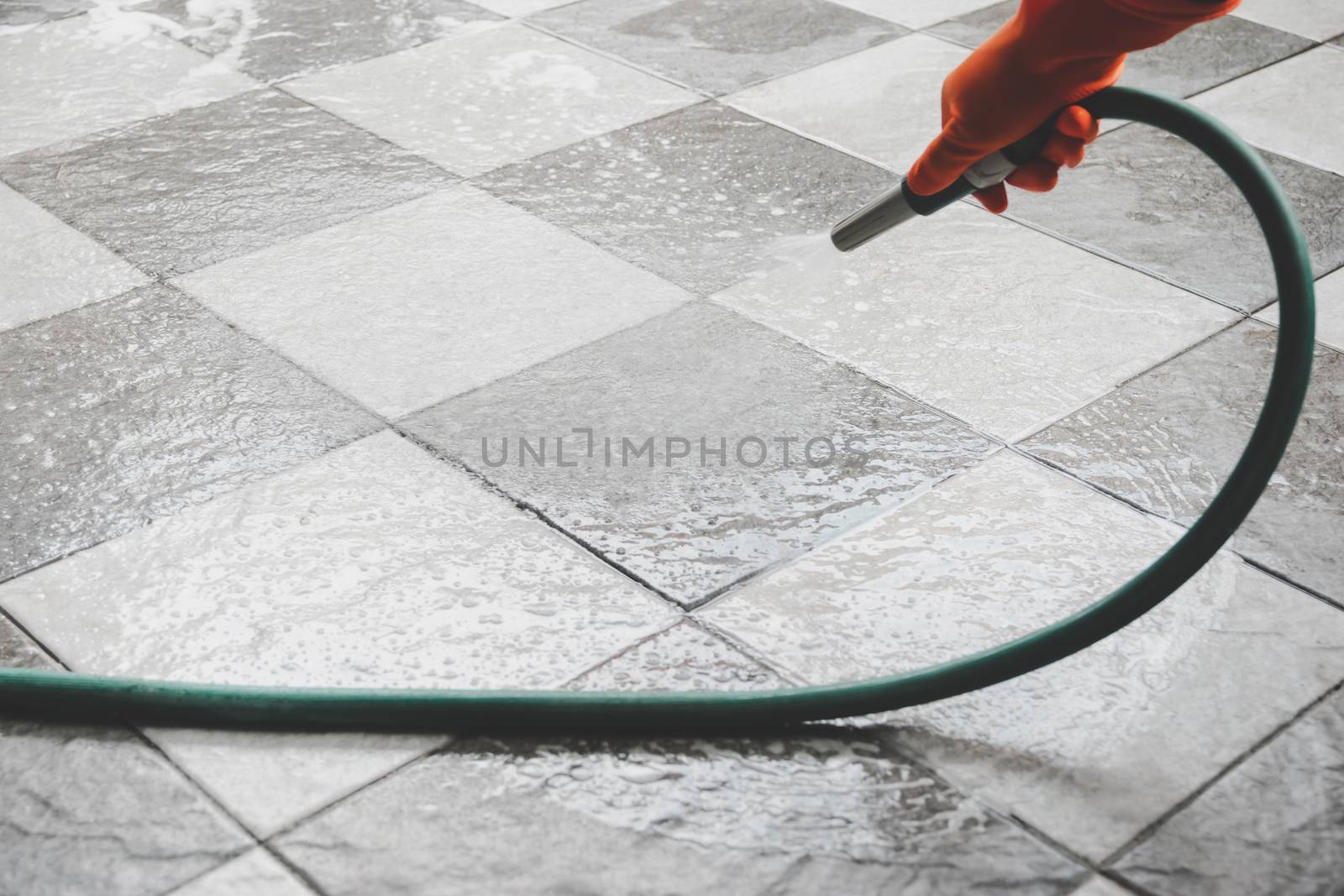 Hand of man wearing orange rubber gloves is use a hose to clean the tile floor.