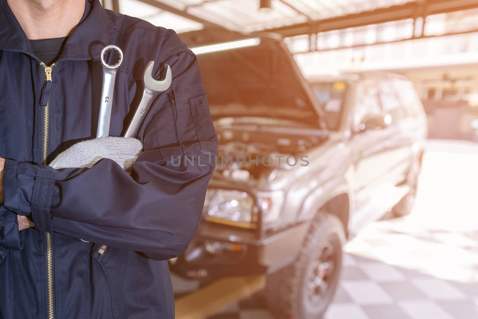 Car repairman wearing a dark blue uniform standing and holding a wrench that is an essential tool for a mechanic and has a backdrop as a car repair center.