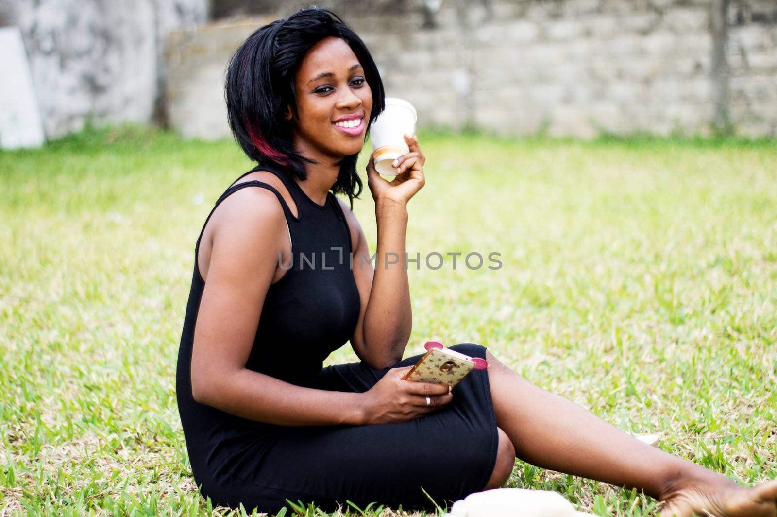 Urban girl in black dress sitting in the garden and holding a cell phone and a cup of coffee