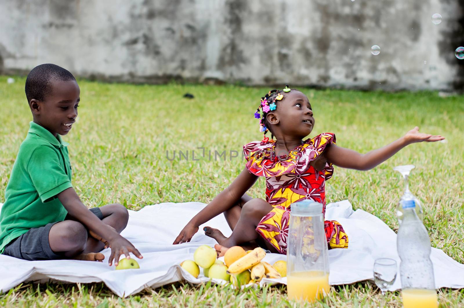 two children sitting on a white tablecloth playing with soap bubbles and fruits during a picnic in a garden